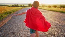 Boy Plays Superhero. Happy little boy are playing superhero. Kid run across road in red cloak at sunset time. Pretty child superhero hero in red cloak in nature. Freedom power concept.