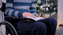 Christian man in wheelchair reading the Bible in front of Christmas tree. Spiritual time and studying the Bible concept. Dolly shot 4k closeup