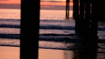 tide washing onto a shore at sunset under a pier 