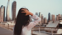 View from back girl walks forward through city Dubai to high-rise buildings skyscrapers on sidewalk next to sea marina. Young woman straightens her luxurious hair with her hand, fluttering in wind.