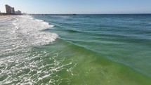 Aerial footage of blue and green ocean waves crashing into a beach going towards a pier.