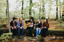 A group of young women sitting on a tree trunk listening to someone play a guitar.