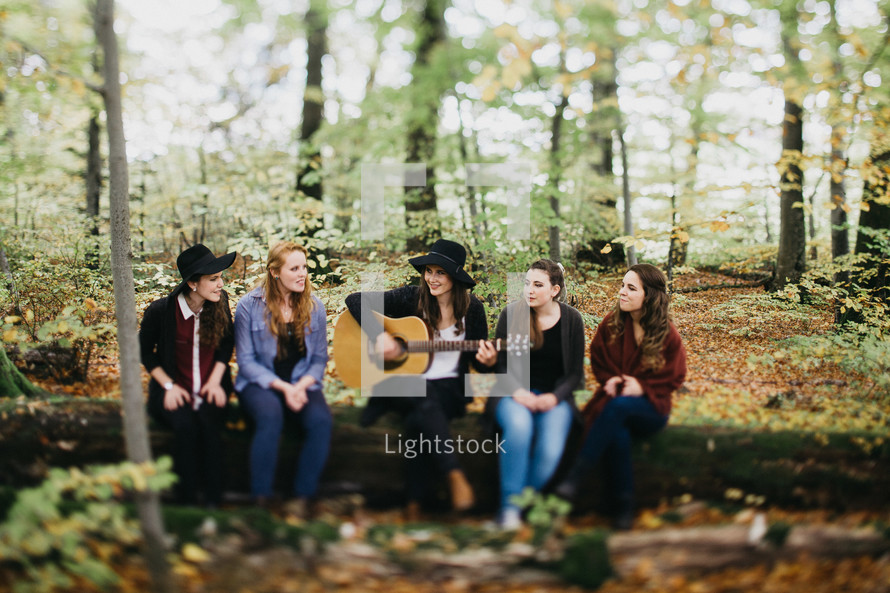 A group of young women sitting on a tree trunk listening to someone play a guitar.