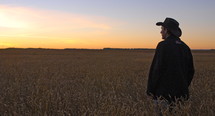 A man wearing a cowboy hat watches the sunrise in a wheat field. A