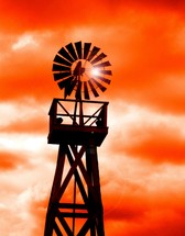 A Windmill silhouetted against an orange sunset  used to harness the power of the wind on a rural farm. 
