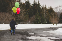 boy child walking in snow carrying a stuffed animal and balloons 