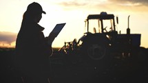 Silhouette woman uses specialized app on a digital tablet PC on background of working tractor in field. Remote control machinery equipment on farmland. Concept optimization of work of small business.