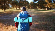 Young male tourist walking with hiking backpack on his shoulders. Walks through the outdoor nature park with accessories for picnic. Mat and equipment. Sporty lifestyle on summer vacation.