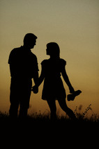 Silhouette of a couple holding hands while standing in a field at sunset.