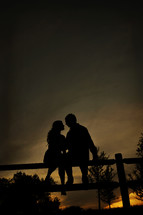 Couple sitting on a fence post embracing at sunset.