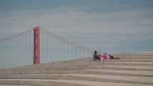 Two people sunbathing leisurely atop the MAAT museum in Lisbon, with the iconic 25th of April Bridge in the background.