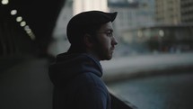 A contemplative young man stands on a bridge looking out towards the city skyline