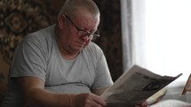 A mature man in glasses reading a newspaper at home. Senior Adult Reading Newspaper Leisure Concept.