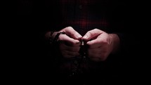 A man's hands wrapped with a wooden Rosary comes out of the dark into the light with praying hands to Jesus.