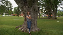 A female college student smiles as she leans on a tree