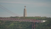 The majestic Santuário de Cristo Rei, or Christ the King monument, stands atop a hill with the iconic 25th of April Bridge in the foreground in Lisbon, Portugal.