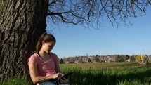A young woman sitting against a tree laughing as she looks at her electronic tablet.