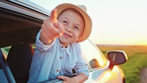 View from car window of child who lean out of window and point his finger a country road on field. Sunny sky, relaxing, enjoying road trip and feeling the air and freedom. Toward adventure, vacation.