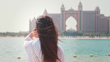 Girl hair beautifully developing in wind in Dubai against of 5 star hotel. Happy woman walking near sea in city. Vacation and sightseeing. Luxury and comfortable tourism season in UAE.