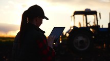 Silhouette woman uses specialized app on a digital tablet PC on background of working tractor in field. Remote control machinery equipment on farmland. Concept optimization of work of small business.
