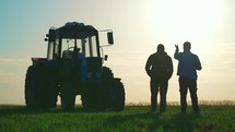 Teamwork concept. Silhouette two male farmers walking in a green field against sunset. Team farmers stand in a field on the background of agricultural machinery. Agronomists discuss harvest.