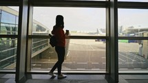 Portrait young woman drinking coffee and standing in waiting room in front of large window with view of parked plane at airport and waiting for her flight. Travel to resort around the world by air.