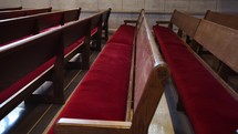 Slow-motion dramatic shot of empty wooden church pews with red fabric to sit on and a brick wall in the background.