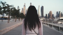 View from back girl walks forward through city Dubai to high-rise buildings skyscrapers on sidewalk next to sea marina. Young woman straightens her luxurious hair with her hand, fluttering in wind.