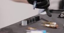 Tattoo Artist Dips The Tip Of The Needle Cartridge Into A Filled Ink Cap On The Table. - close up shot