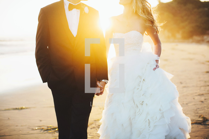 torso of a bride and groom holding hands walking on a beach