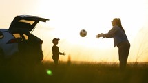 Mom and baby son in nature playing ball in field with tall grass against the back of sunset and car. Family spends time weekend in walk a outdoor in the park outside the city. People enjoying summer.