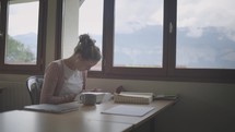Young woman reads and studies the Bible at a table inside a room with large windows and natural light