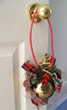 A big brass Jingle Bells with a red ribbon and red and green holly leaves adorn a brass door knob doorway entrance to a home's interior bringing out color, warmth and cheer for the Christmas holiday season.