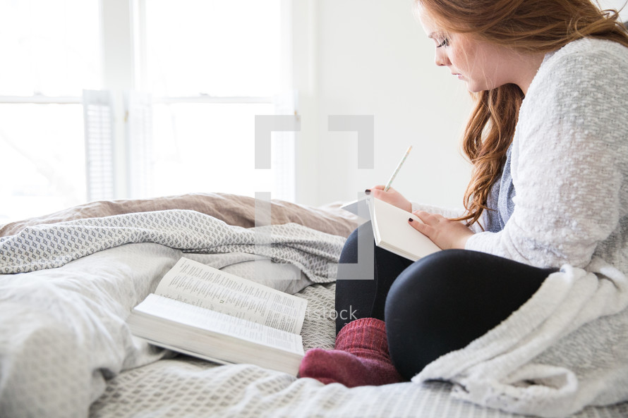 young woman sitting in bed reading a Bible and writing in a journal 