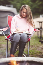woman reading a Bible and taking notes in front of a fire pit 