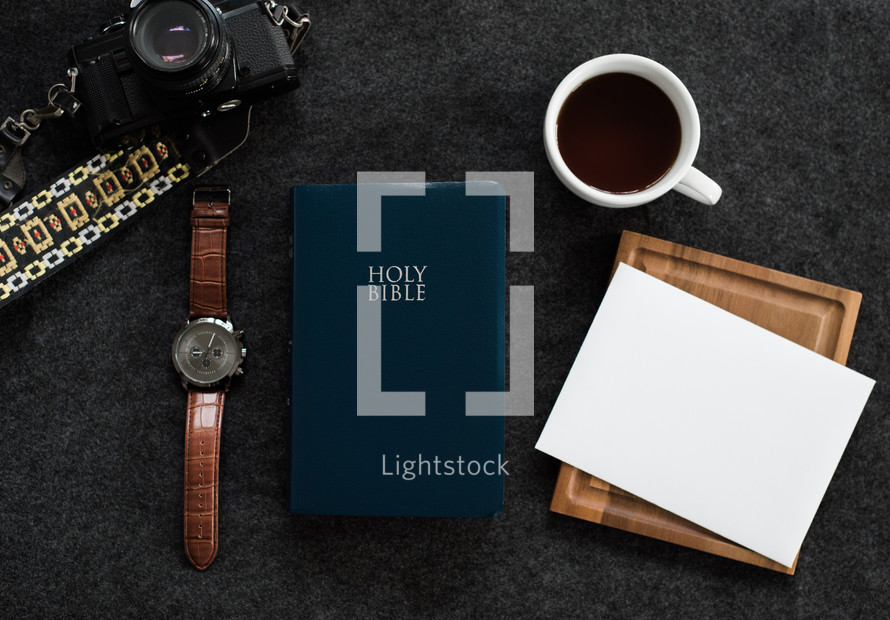 A Bible, wristwatch, cup of coffee, camera and a blank envelope on a black background.