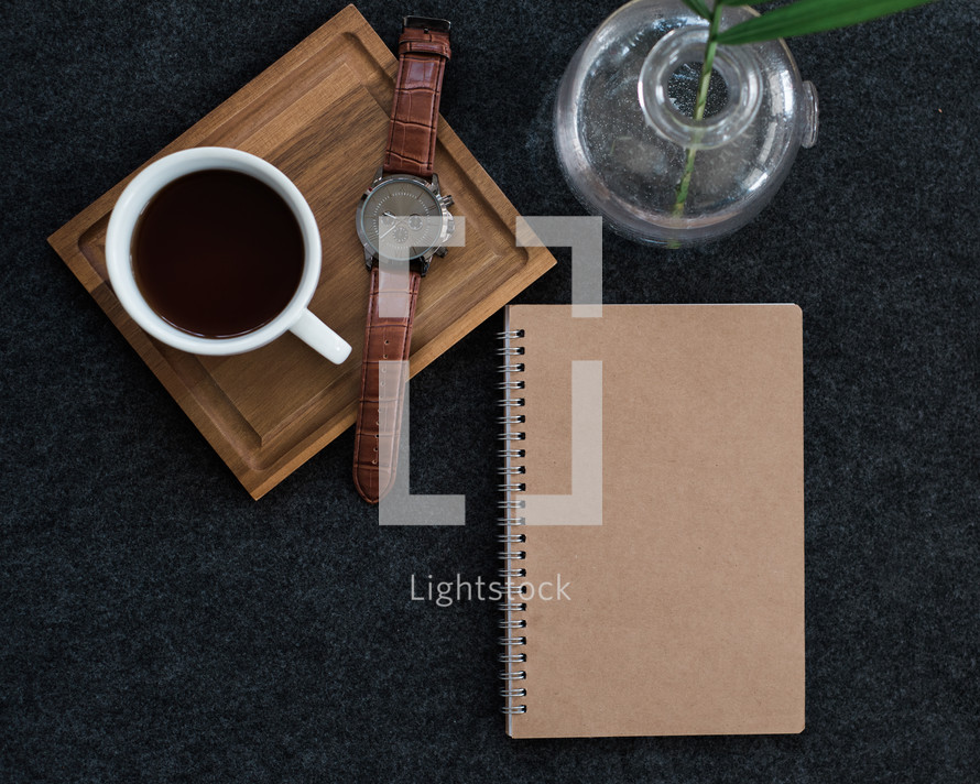 A wristwatch and cup of coffee on a wooden board next to a spiral notebook and a plant in a vase.