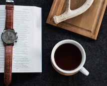 A wristwatch laying on an open Bible next to a cup of coffee and an antler.