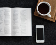 An open Bible next to a cell phone and a cup of coffee.