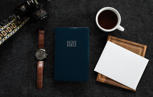 A Bible, wristwatch, camera, cup of coffee and a blank envelope on a black background.