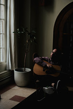a man sitting in a chair by a window holding a guitar 