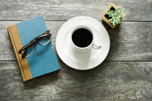 reading glasses, book, coffee cup, saucer, and house plant on a table 