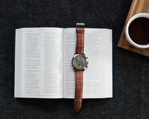 A wristwatch laying on an open Bible and a cup of coffee on a black background.