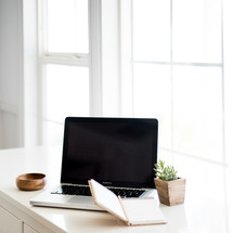 A laptop computer and notebook on a white surface by a windowsill.