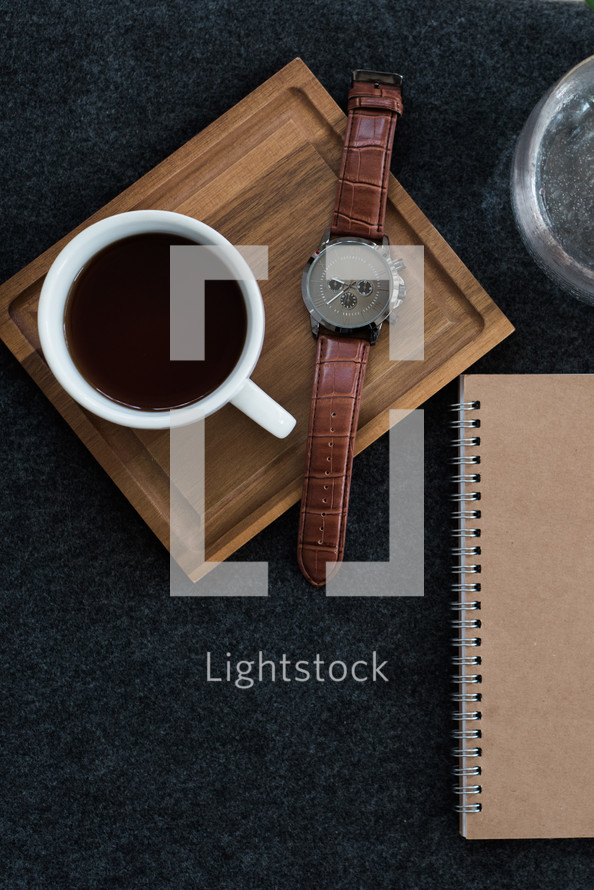 A cup of coffee and wristwatch on a wooden coaster next to a vase and a spiral notebook.