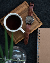 A wristwatch and a cup of coffee next to a spiral notebook and a plant in a vase.