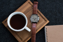 A cup of coffee and wristwatch on a wooden board next to a spiral notebook.