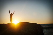 silhouette of a man standing with raised arms on a rock near the ocean 