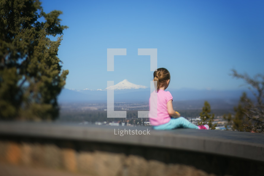 a girl sitting on a wall looking out at a snow capped mountain peak 