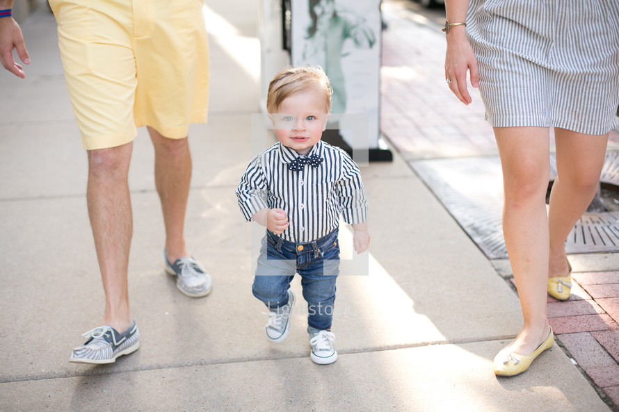 Toddler boy in a bow tie walking on a sidewalk with his parents.